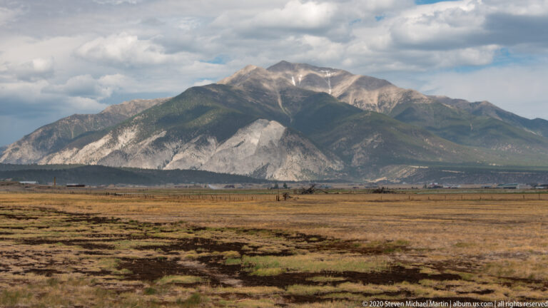 A broad dry valley and fields lead toward a forested mountain range with pale exposed rock beneath broken clouds.