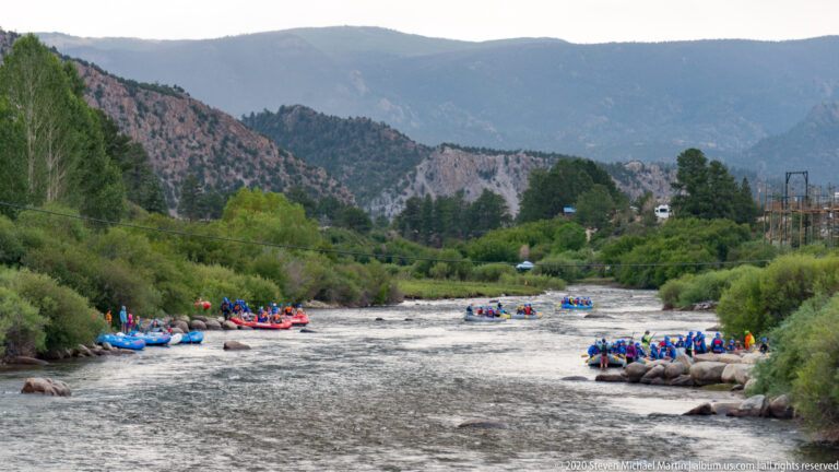 Colorful rafts carrying groups of boaters move through a rocky river channel between wooded mountain slopes.