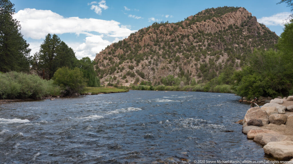 The Arkansas River flows past a boulder-lined bank beneath a steep, pine-covered rocky hillside.