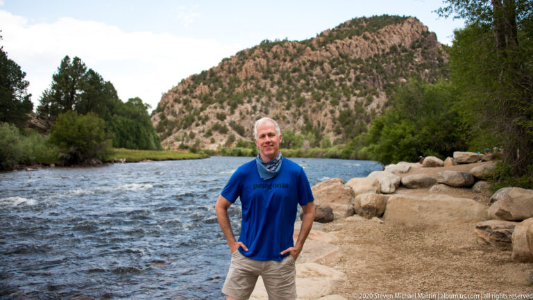 A man in a blue shirt stands beside the Arkansas River, with boulders, wooded slopes, and distant visitors behind him.
