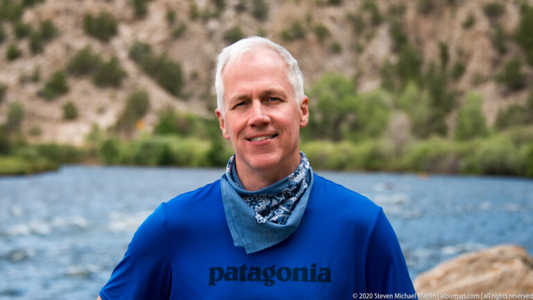 A smiling man in a blue shirt and patterned neck gaiter stands before a softly blurred river and green bank.
