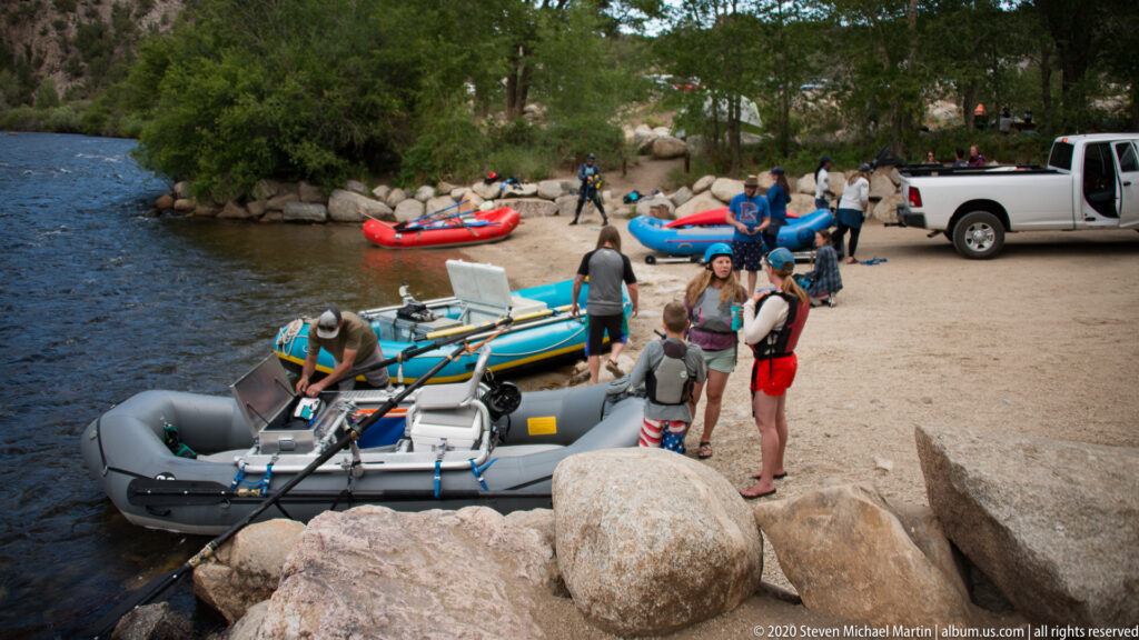 Visitors load and prepare inflatable rafts on a sandy riverbank beside a pickup truck and boulders.