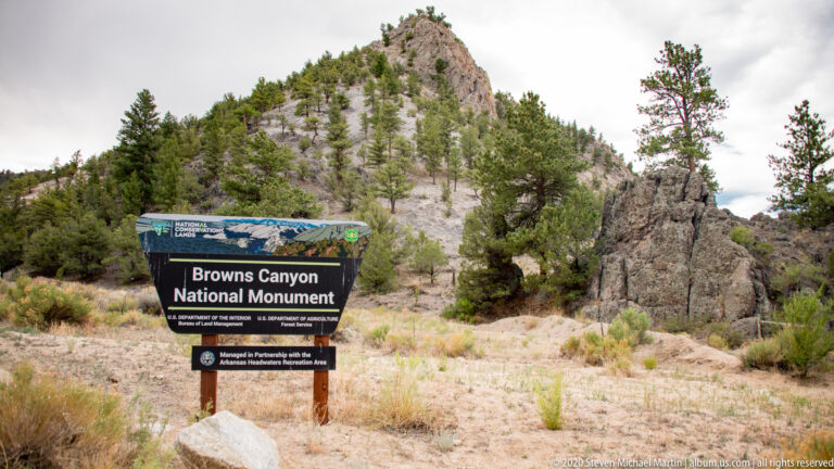 A Browns Canyon National Monument sign stands below a rocky pine-covered hill amid pale grasses and shrubs.