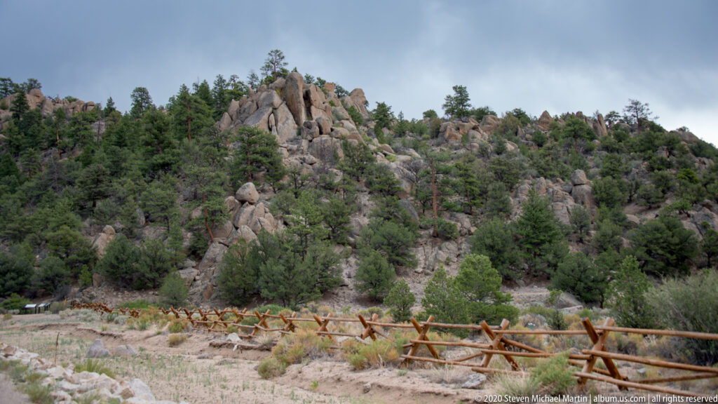 A split-rail wooden fence crosses dry ground below a rocky hillside dotted with pine trees.