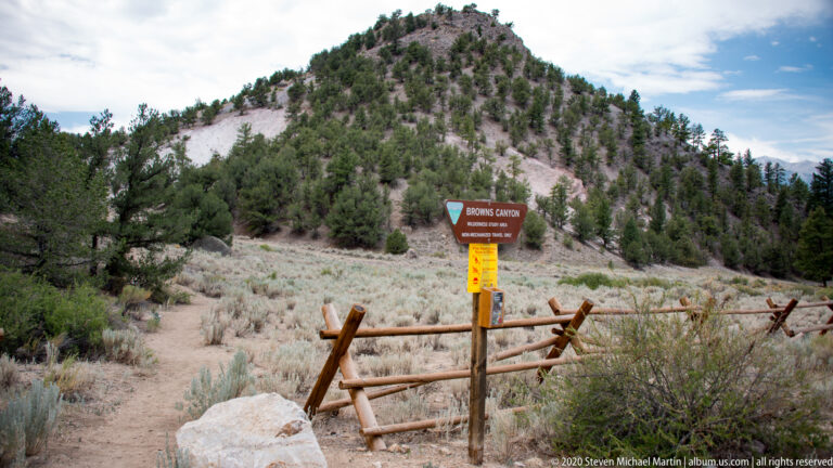 A Browns Canyon Wilderness Study Area sign marks a path through a wooden fence toward a rocky, pine-dotted hill.