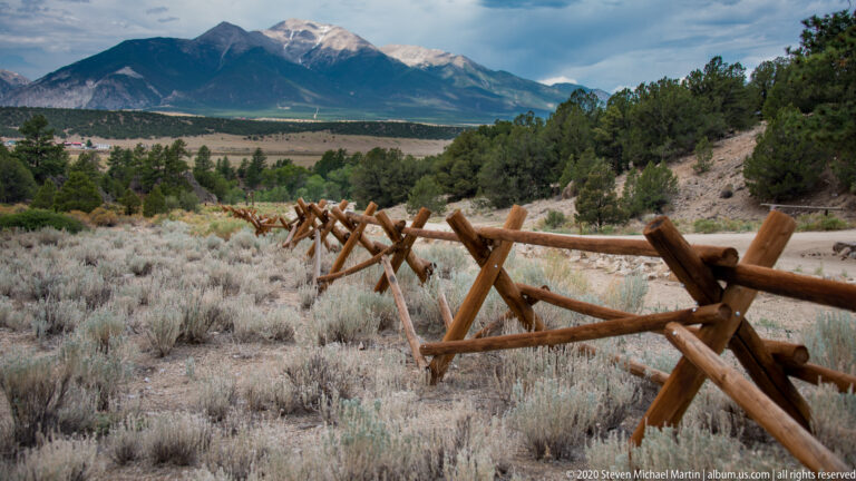 A split-rail fence recedes across dry grassland toward a broad valley and distant mountain ridge.