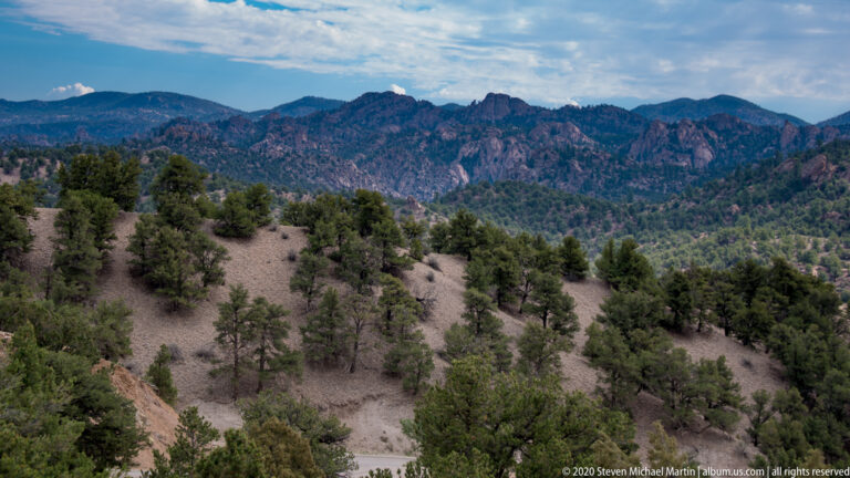 Scattered pines cover a hillside below a rugged, cloud-shadowed mountain ridge in Colorado.