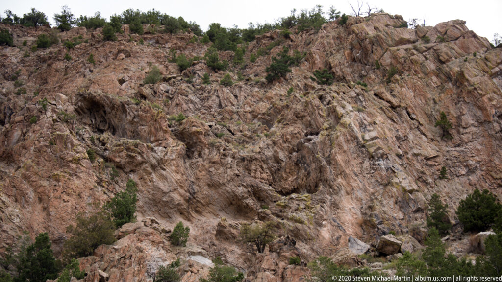 Fractured pink and gray rock slabs form a steep outcrop with sparse shrubs and small pines along the ledges.