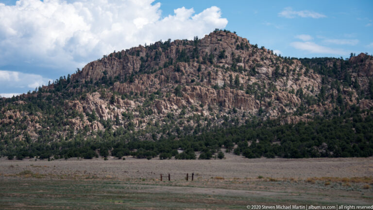 A rocky, pine-covered ridge rises beyond a broad valley grassland beneath bright cumulus clouds.