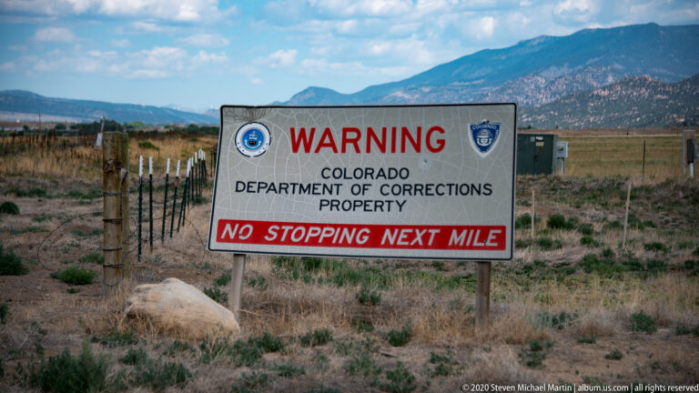 A Colorado Department of Corrections sign warns “No Stopping Next Mile” beside a fenced field with mountains behind.