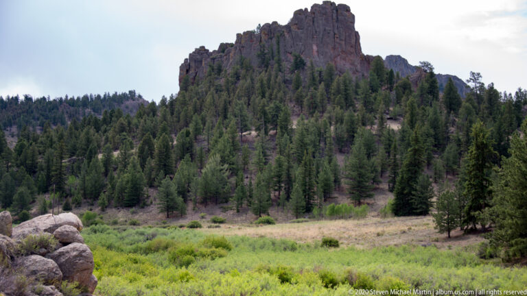 Dark granite pinnacles rise above a pine-covered slope and green meadow, with rounded boulders in the foreground.