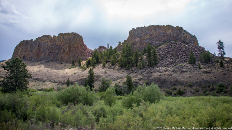 Two rounded rocky ridges separated by a low saddle above shrubs and scattered conifers.