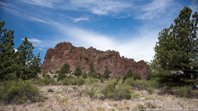 Red-brown rock wall beyond scrub and pines, with a sandy path in the foreground.