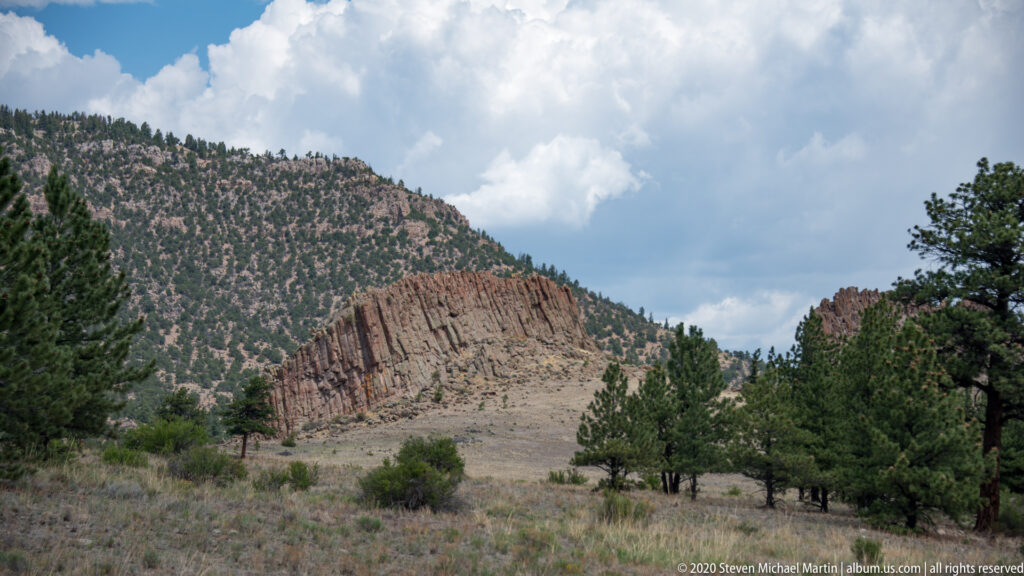 Red-brown cliff across a mountain valley, framed by pine-covered slopes and cloudy sky.