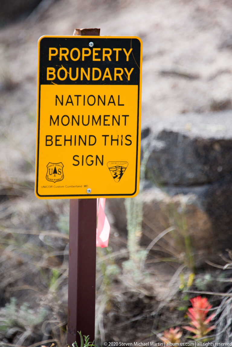 A yellow boundary sign says the national monument begins behind it and displays agency emblems.
