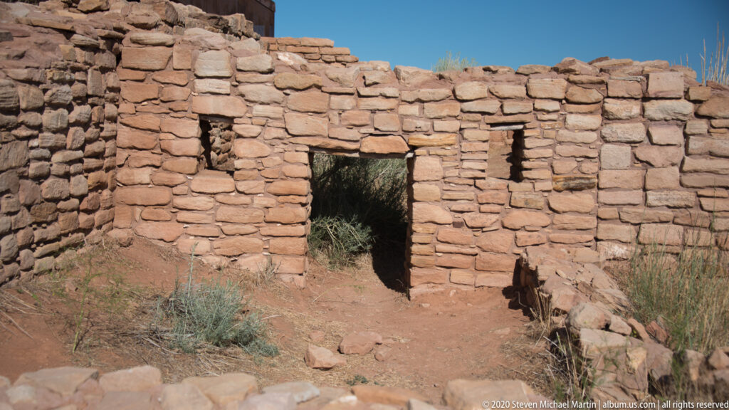 A narrow path leads to a dark doorway in a thick pueblo wall.