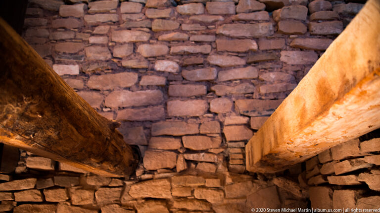 Two timber beams project across a dim stone chamber toward a rough masonry wall.