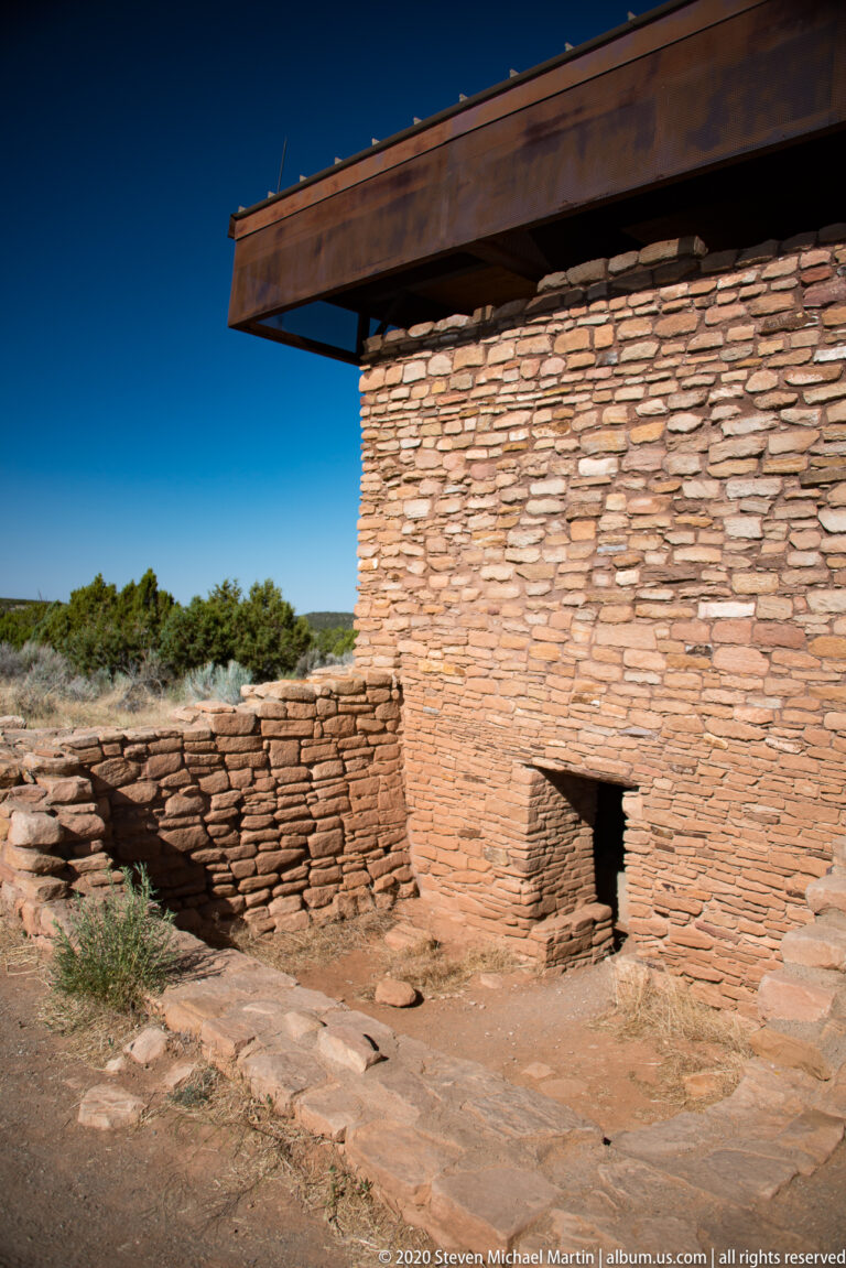 A roofed stone structure with a low doorway stands beside a short masonry wall.
