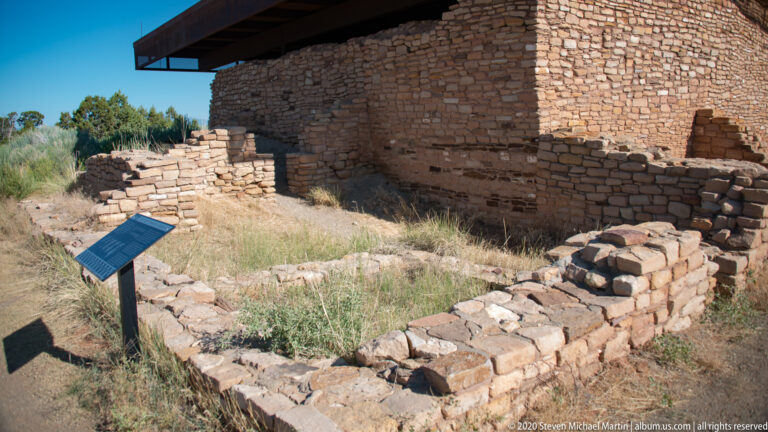 Low stone foundations with an interpretive marker lie beside a taller roofed structure.