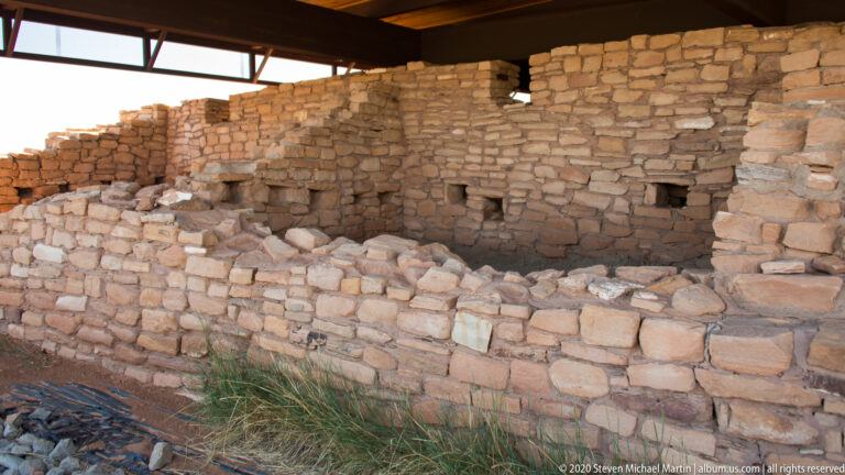 Masonry rooms with small wall openings sit beneath a protective roof at Lowry Pueblo.