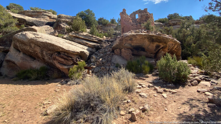 Sandstone boulders and low pueblo walls sit among juniper and dry grasses.