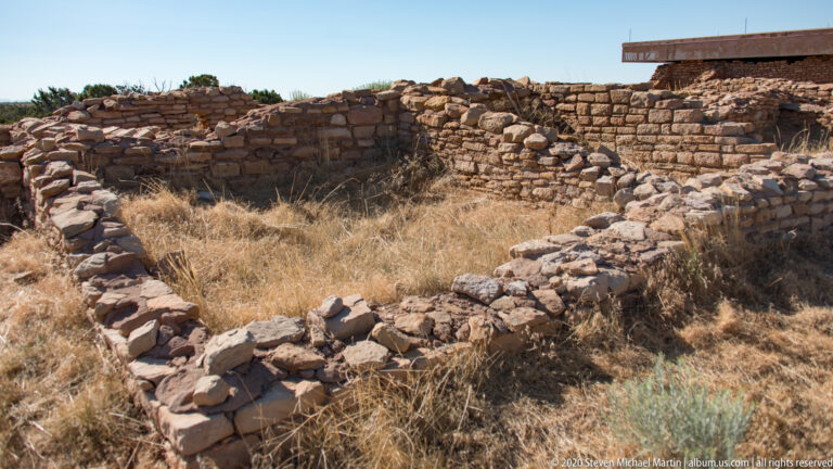 Low stone walls curve around a grass-filled enclosure beside taller ruins at Lowry Pueblo.