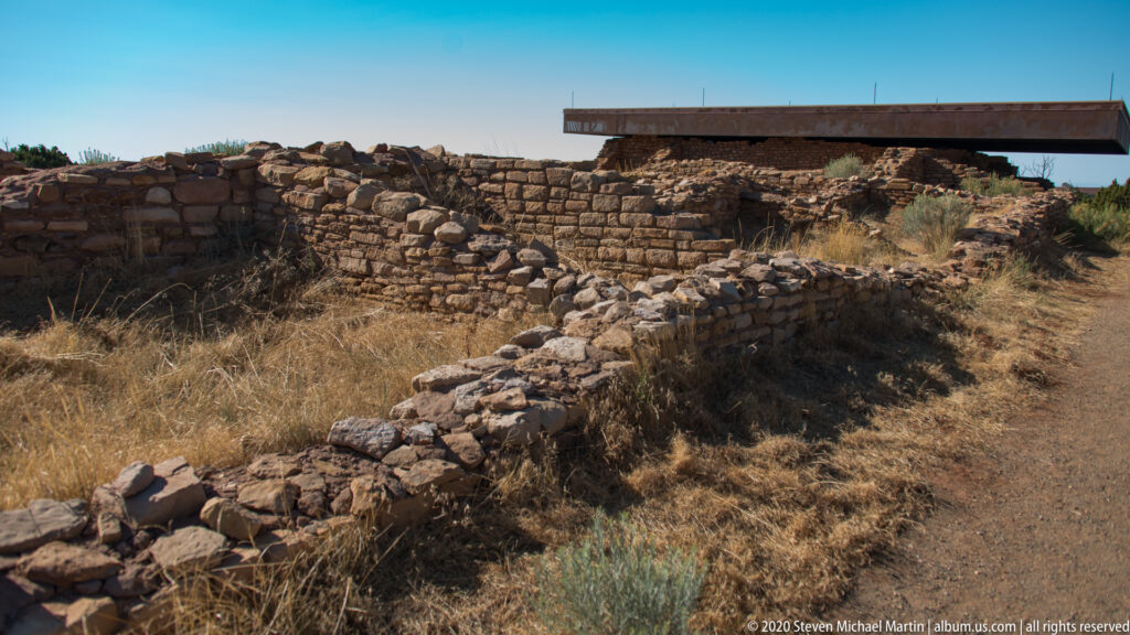 Low foundations lead toward taller stone walls beneath a protective canopy at Lowry Pueblo.