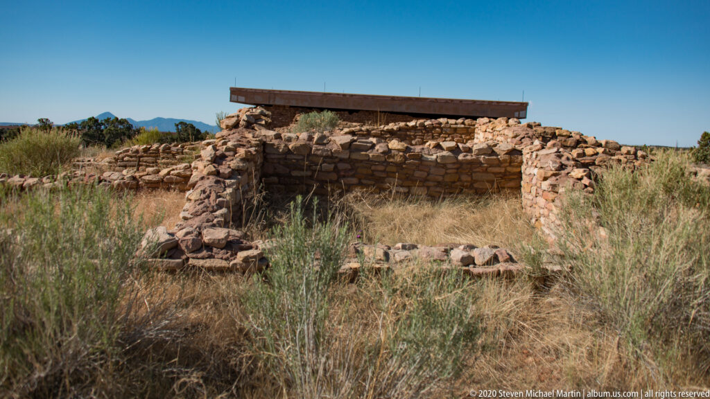 A flat roof shades a long stone wall and small opening above dry grass.