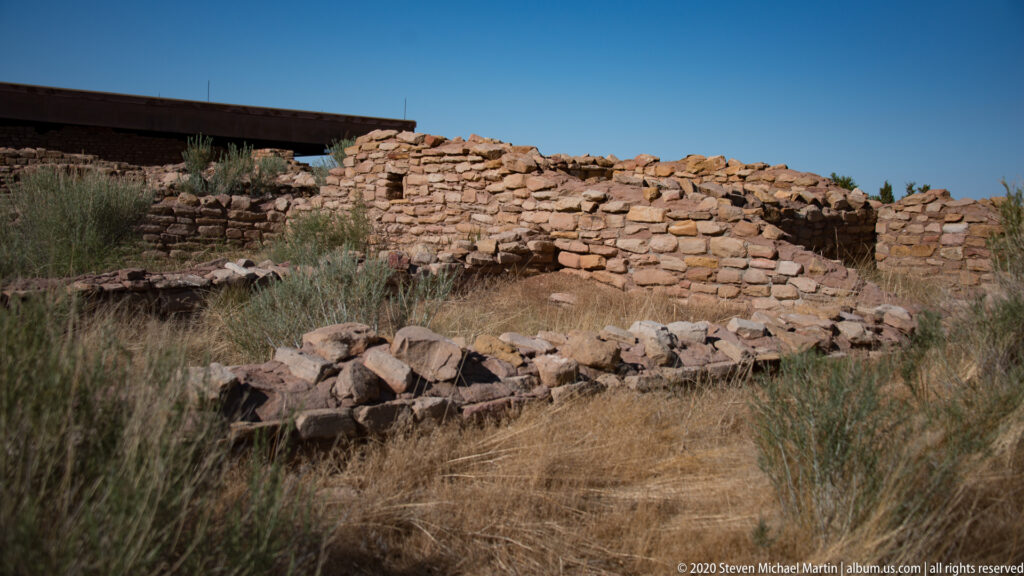A low stone wall and loose masonry sit beneath a broad canopy among dry grasses.