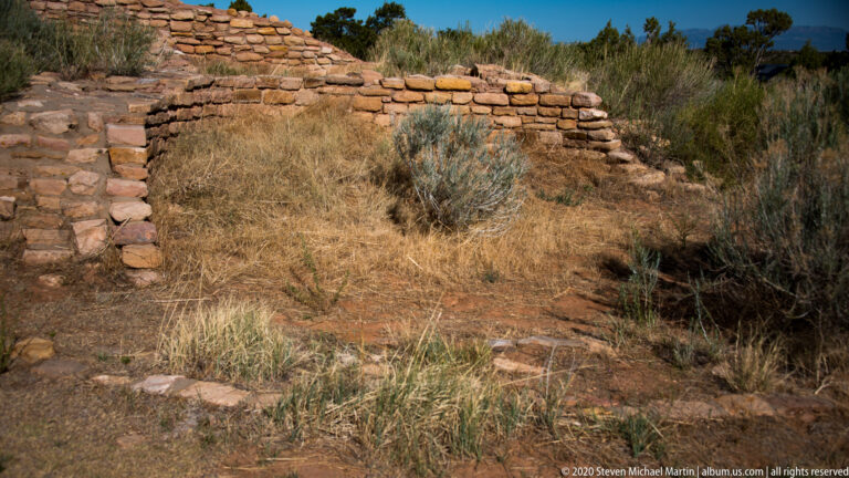 A curved pueblo wall borders dry ground and grasses, with shrubs behind it.