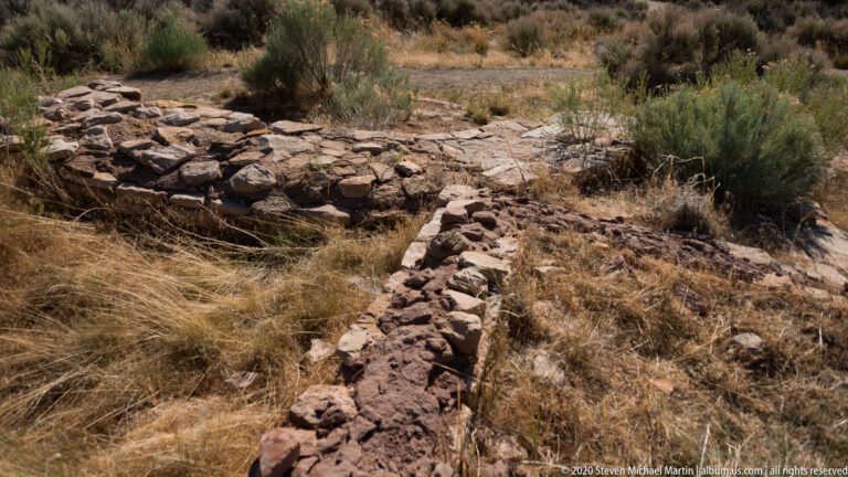 A broken stone foundation runs diagonally through dry grass and shrubs at Lowry Pueblo.