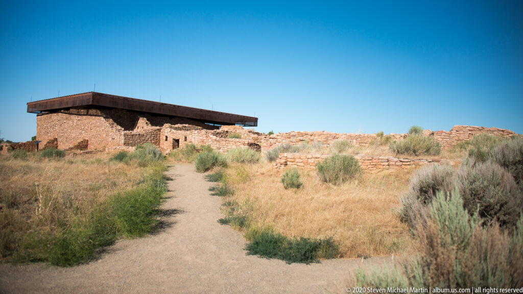A dirt path leads to a long roofed masonry structure surrounded by grass and stone walls.