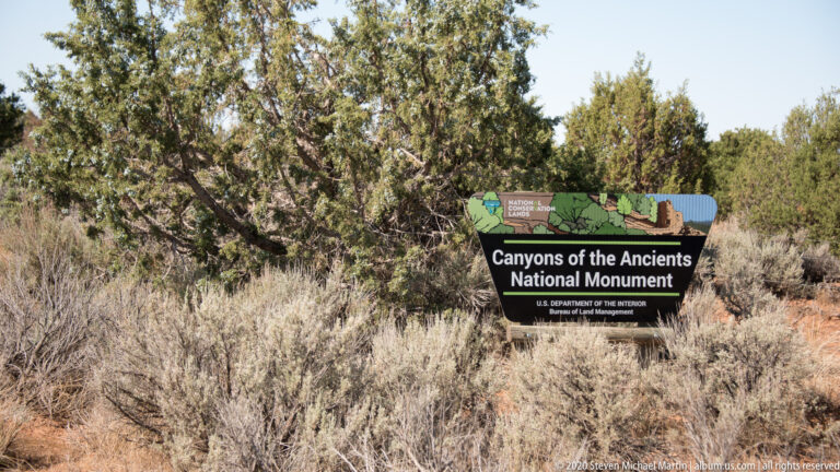 A black Canyons of the Ancients National Monument sign stands among sagebrush and juniper.