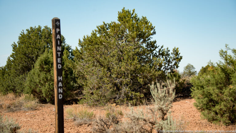 A dark vertical sign marked Painted Hand stands among juniper trees and dry brush.