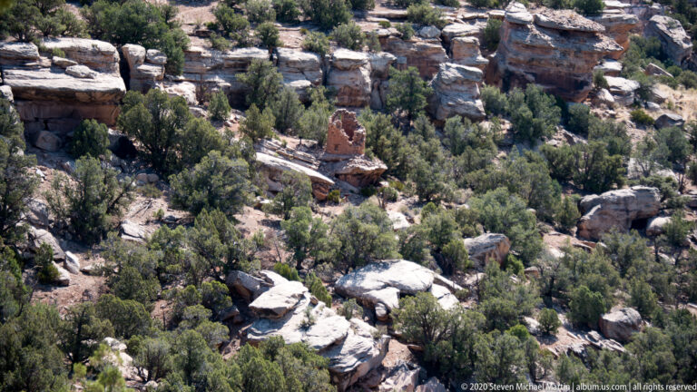 A small reddish masonry ruin is surrounded by pinyon-juniper trees and pale sandstone ledges.