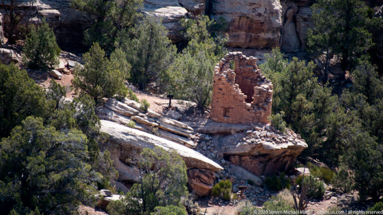A reddish masonry tower ruin stands on a sandstone shelf among junipers, with a person below for scale.