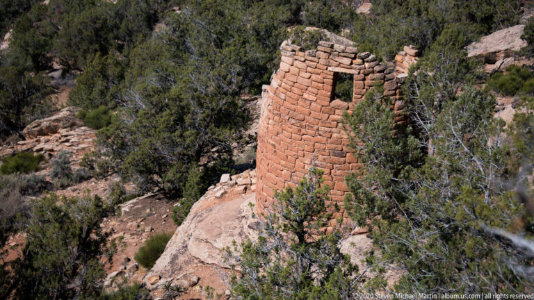 A cylindrical stone tower ruin with a rectangular opening stands on sandstone among junipers.