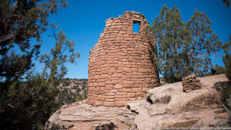 A round masonry tower with a rectangular opening stands on a sandstone ledge among junipers.