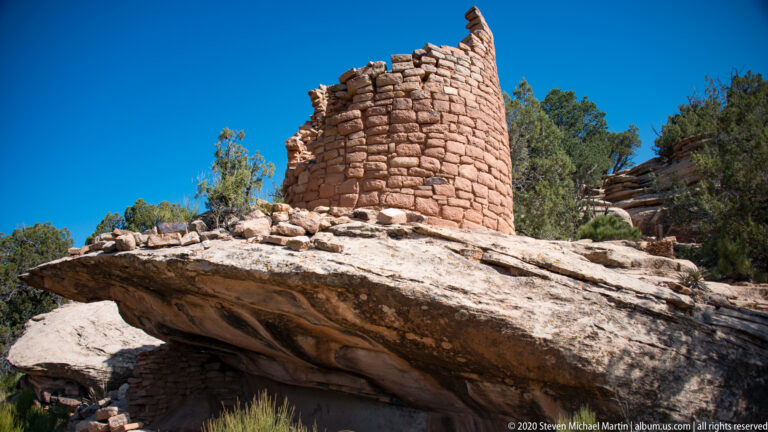 A broken masonry tower stands on a broad projecting sandstone ledge above a dark recess.