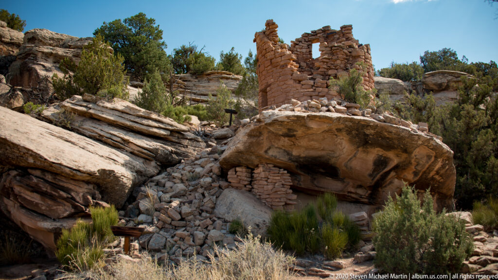 A broken reddish tower with a dark opening stands among sandstone shelves, rubble, and junipers.