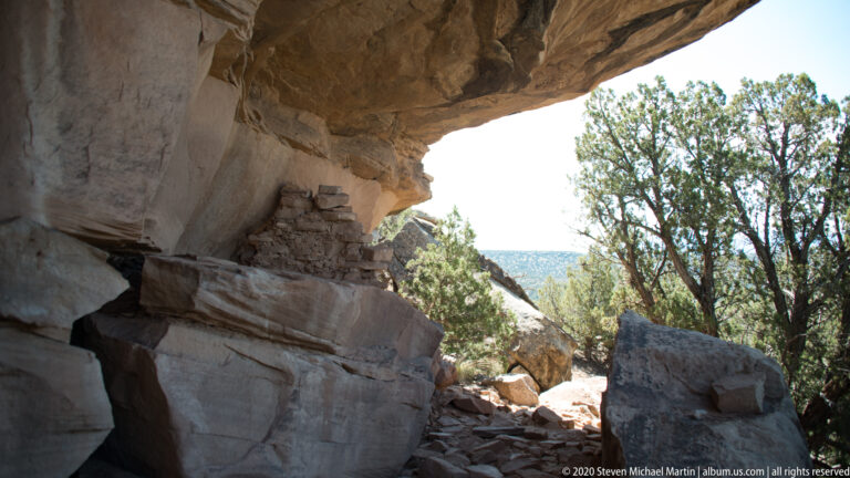 A shaded sandstone overhang shelters broken masonry beside a bright opening and tree-covered slope.