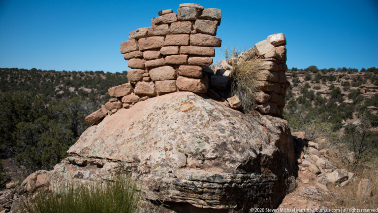 A reddish masonry wall stands atop a rounded boulder, beside a broken wall and faint orange marks.