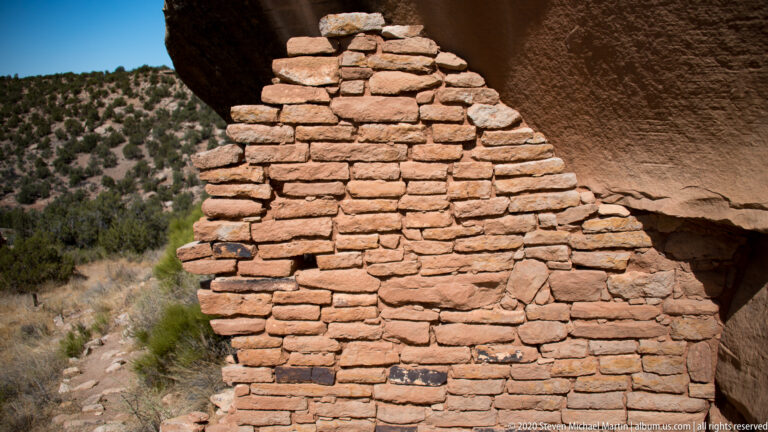 A rough stone wall stands beneath a sandstone overhang, with junipers visible through an opening.