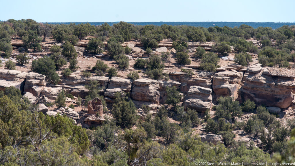 A small reddish tower ruin appears among junipers and pale sandstone outcrops across a wooded plateau.
