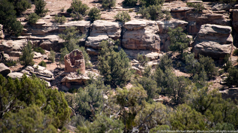 A distant reddish tower ruin stands among junipers and sandstone ledges on a wooded slope.