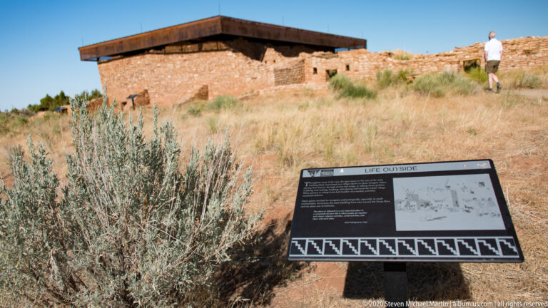 A visitor approaches a roofed stone building beyond a “Life Outside” interpretive sign.