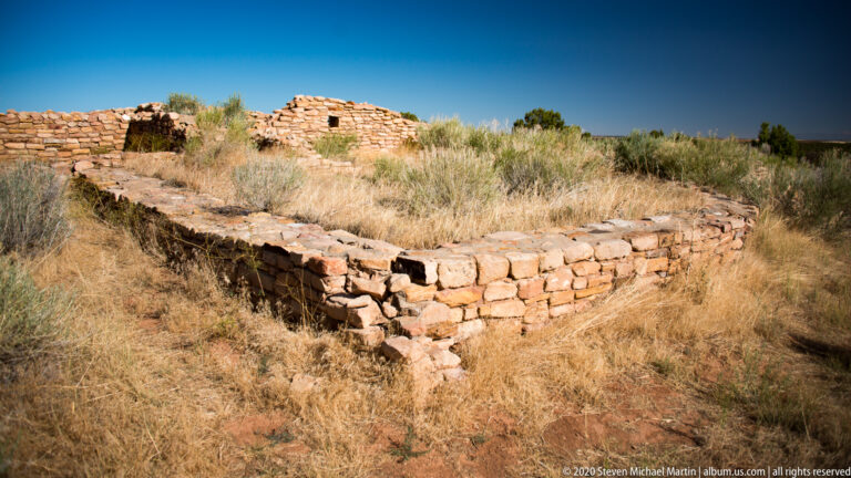 A curved stone wall encloses dry grass beneath a clear blue sky.