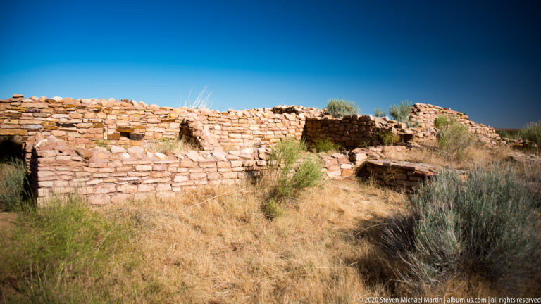 Low pueblo walls and a dark opening extend across a dry grassy site.