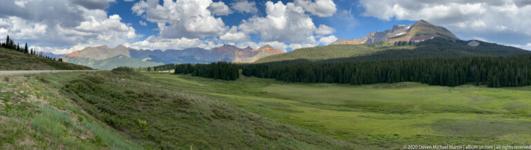 A pointed mountain peak rises over a forested valley as a road curves along the left slope.
