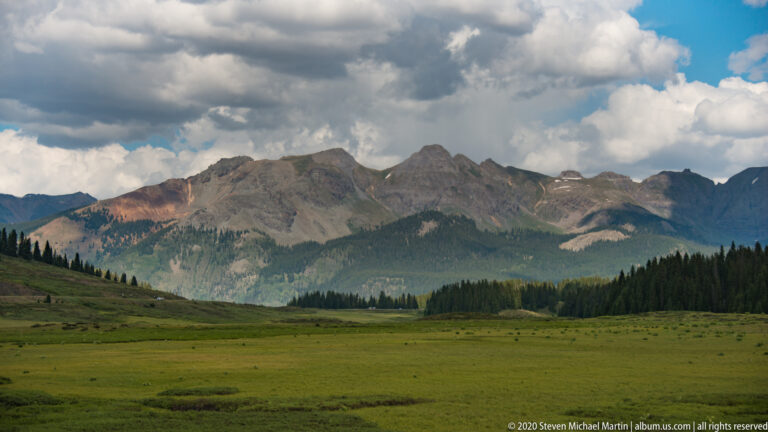 A green alpine meadow and conifer forest extend toward a dark ridge under heavy clouds.